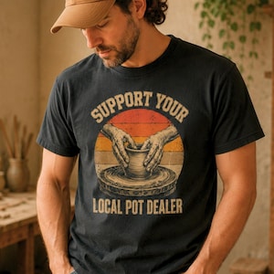 May include: A black short-sleeve t-shirt featuring a graphic of hands shaping clay on a pottery wheel set against a sunset-coloured circle. The text "SUPPORT YOUR LOCAL POT DEALER" is printed in a curved font above and below the central illustration.