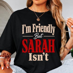 May include: A black short-sleeved t-shirt featuring the distressed white text I'm Friendly But and the word SARAH in large bold red letters, followed by the white text Isn't. The design is centered on the chest of the garment.