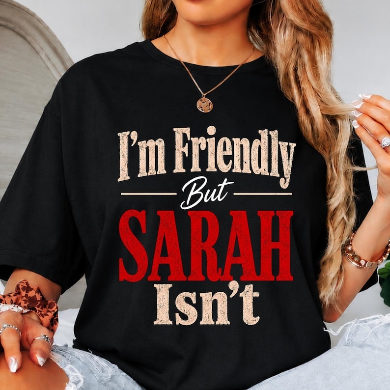 May include: A black short-sleeved t-shirt featuring the distressed white text I'm Friendly But and the word SARAH in large bold red letters, followed by the white text Isn't. The design is centered on the chest of the garment.