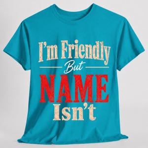 May include: A bright turquoise short-sleeve t-shirt featuring the text "I'm Friendly But NAME Isn't" printed on the front. The words "I'm Friendly But" and "Isn't" are in a distressed beige font, while the word "NAME" is in large red letters.