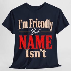 May include: A dark navy blue short-sleeve t-shirt featuring the text "I'm Friendly But NAME Isn't" printed on the front. The words "I'm Friendly But" and "Isn't" are in a distressed beige font, while the word "NAME" is in large red letters.
