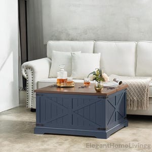 May include: A navy blue wooden coffee table with a brown top, featuring a farmhouse style design. The table has an X-shaped design on the front and sides. A decanter, glasses, and flowers are on top of the table.