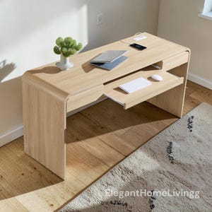 May include: A light oak wood desk with rounded side panels, featuring a pull-out keyboard tray, a flat work surface, and a small drawer. On the desk sits a silver laptop, a white computer mouse, a white keyboard, and a small green plant.