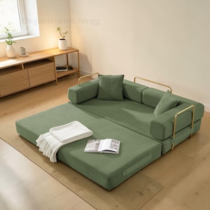 May include: A modular green sofa with gold-colored metal accents. The sofa is partially unfolded, revealing a pull-out section. A white blanket and an open book rest on the unfolded portion. The text "ElegantHomeLivingg" is visible.