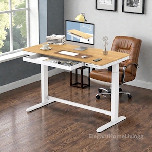 May include: A light wood-tone adjustable height desk with a white frame and a built-in drawer. The desk has a computer monitor, a brown leather desk chair, and a small side table. The desk is in a room with a window and hardwood floors.