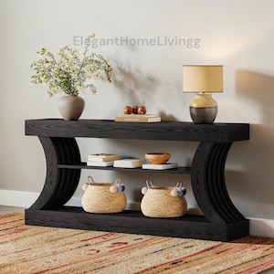 May include: A black console table with curved sides, two shelves, and a lower base. The table is decorated with a vase of flowers, a lamp, books, a bowl, and two woven baskets with pom-poms. A rug is beneath the table.