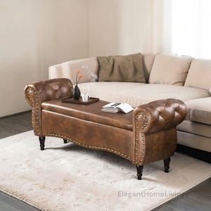 May include: A brown leather storage bench with rolled arms and button tufting. The bench has nailhead trim and dark wooden legs. A tray with a cup and vase sits on top, with a book nearby. The bench is in front of a beige sofa.