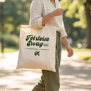 May include: A canvas tote bag with the text "Tortoise Swag" in green script and a green turtle graphic. The bag is carried by a person walking on a path in a park. The bag is a natural beige color.