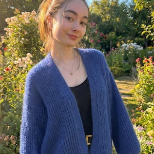 K&ouml;nnte beinhalten: Ein blauer Cardigan mit langen &Auml;rmeln. Der Cardigan wird &uuml;ber einem schwarzen Oberteil und einem goldenen G&uuml;rtel getragen. Der Hintergrund zeigt einen Garten mit Blumen und Gr&uuml;n, und eine Person tr&auml;gt den Cardigan.