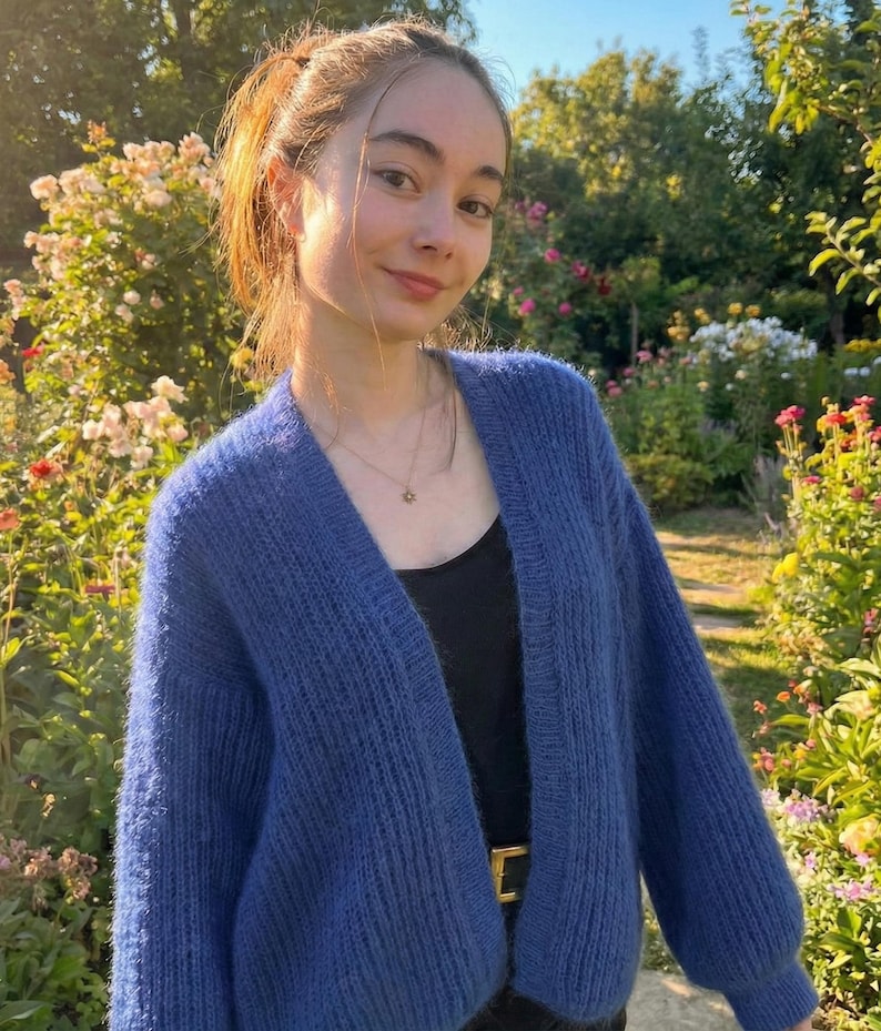 K&ouml;nnte beinhalten: Ein blauer Cardigan mit langen &Auml;rmeln. Der Cardigan wird &uuml;ber einem schwarzen Oberteil und einem goldenen G&uuml;rtel getragen. Der Hintergrund zeigt einen Garten mit Blumen und Gr&uuml;n, und eine Person tr&auml;gt den Cardigan.