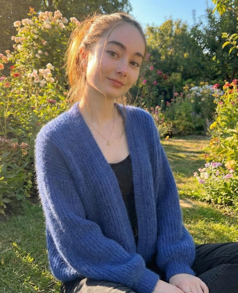 K&ouml;nnte beinhalten: Ein blauer Cardigan mit V-Ausschnitt und langen &Auml;rmeln. Der Cardigan ist aus einem weichen, strukturierten Material gefertigt. Die Person tr&auml;gt ein schwarzes Oberteil und sitzt im Gras in einem Blumengarten.