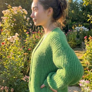 May include: A bright green, chunky knit cardigan. The sweater has a V-neck and long sleeves. The person is standing in a garden with flowers and greenery in the background.