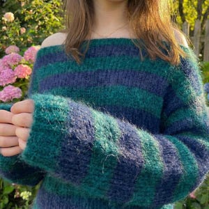 May include: A cozy off-the-shoulder sweater in shades of teal and navy blue. The sweater features horizontal stripes and long sleeves. The knitwear appears soft and fluffy, perfect for cooler weather. The style is casual and comfortable.