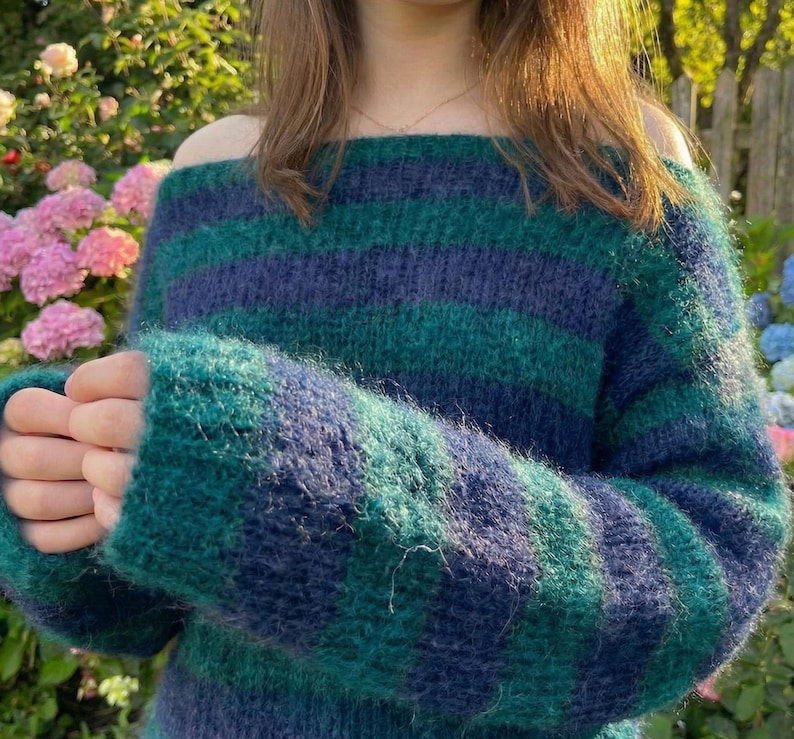 May include: A cozy off-the-shoulder sweater in shades of teal and navy blue. The sweater features horizontal stripes and long sleeves. The knitwear appears soft and fluffy, perfect for cooler weather. The style is casual and comfortable.