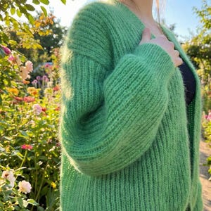 May include: A vibrant green, open-front cardigan made of a soft, textured knit. The long-sleeved sweater has a relaxed fit and is shown outdoors with a garden background. The cardigan is a solid color and has a V-neck.