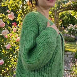 May include: A vibrant green, chunky knit cardigan. The sweater has a ribbed texture and a relaxed fit. The image is set outdoors with a garden background, featuring pink roses and greenery. The cardigan is a solid color and appears soft and cozy.