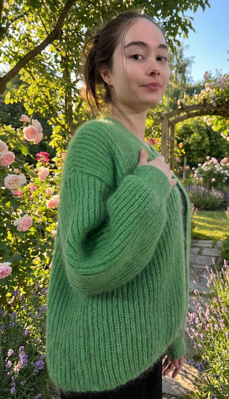 May include: A vibrant green, chunky knit cardigan. The sweater has a ribbed texture and a relaxed fit. The image is set outdoors with a garden background, featuring pink roses and greenery. The cardigan is a solid color and appears soft and cozy.