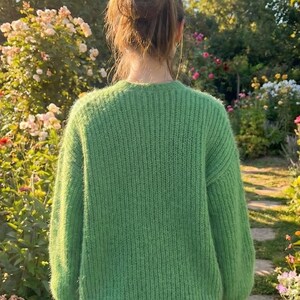 May include: A person wearing a bright green, textured knit sweater. The sweater has a crew neck and long sleeves. The person is standing in a garden with a stone path and colorful flowers.