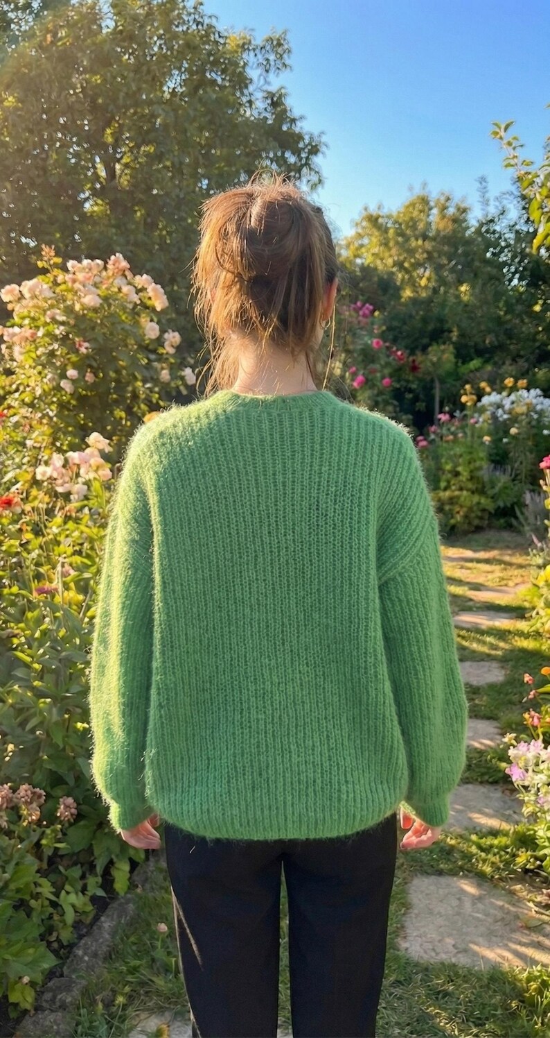 May include: A person wearing a bright green, textured knit sweater. The sweater has a crew neck and long sleeves. The person is standing in a garden with a stone path and colorful flowers.