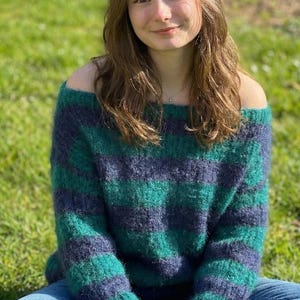 May include: A green and navy blue striped off-the-shoulder sweater. The sweater is made of a fuzzy material and is paired with blue jeans. The person is sitting on green grass.