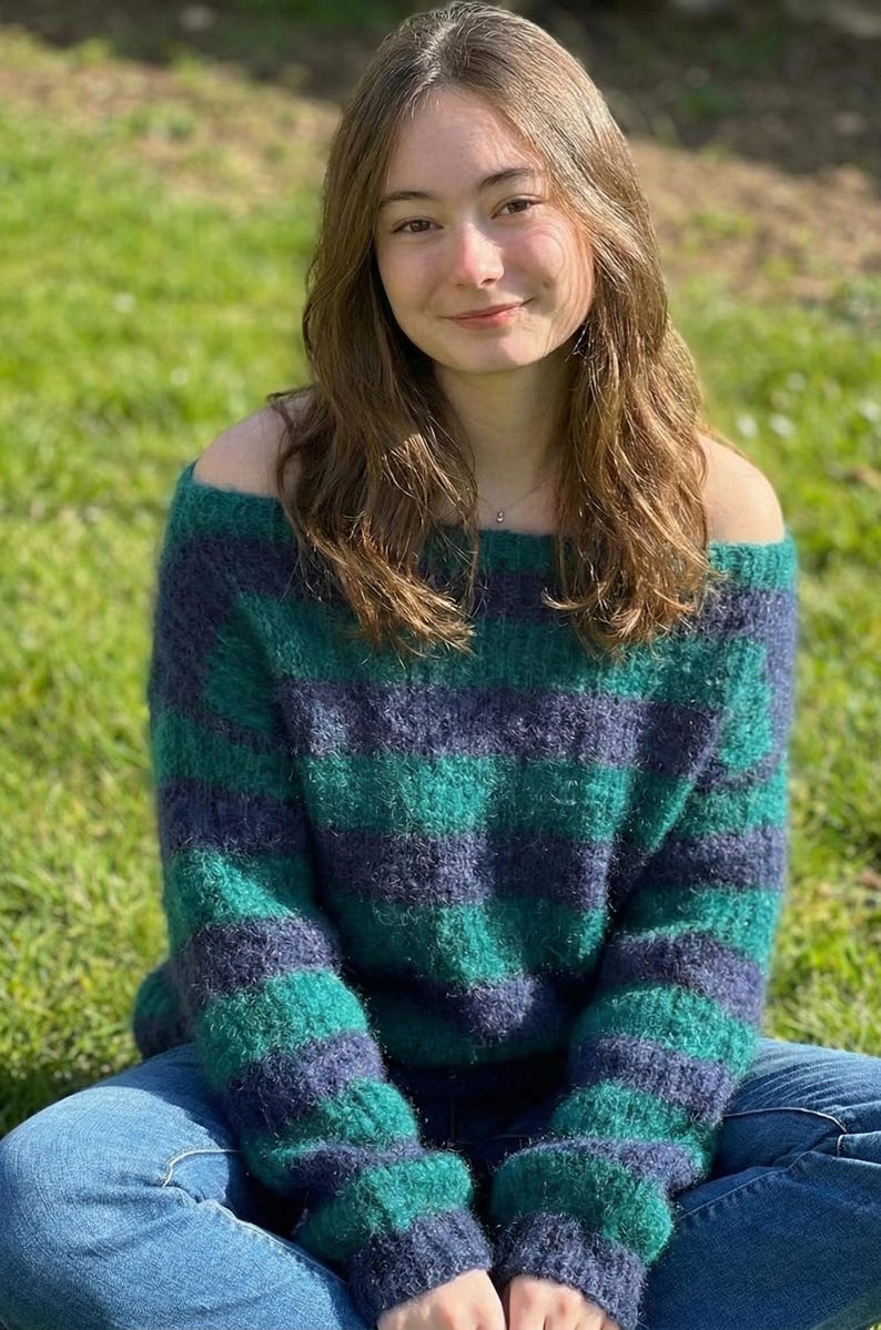 May include: A green and navy blue striped off-the-shoulder sweater. The sweater is made of a fuzzy material and is paired with blue jeans. The person is sitting on green grass.