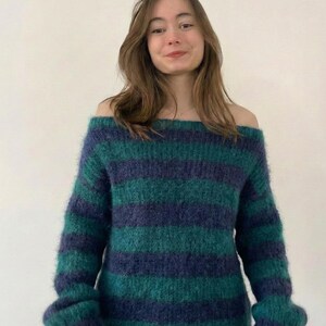 May include: Off-the-shoulder sweater with horizontal stripes in shades of teal and navy blue. The sweater has long sleeves and a soft, textured appearance. The model is wearing the sweater.