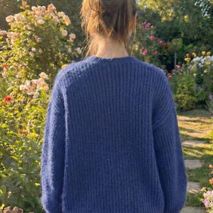 K&ouml;nnte beinhalten: Eine Person tr&auml;gt einen marineblauen Strickpullover mit Rundhalsausschnitt. Der Pullover hat ein strukturiertes Aussehen und besteht aus einem weichen Material. Der Hintergrund zeigt einen Garten mit Blumen und Gr&uuml;n.