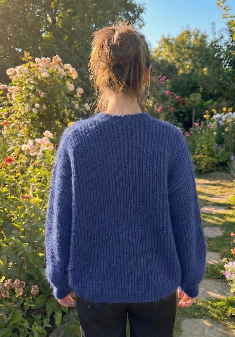 K&ouml;nnte beinhalten: Eine Person tr&auml;gt einen marineblauen Strickpullover mit Rundhalsausschnitt. Der Pullover hat ein strukturiertes Aussehen und besteht aus einem weichen Material. Der Hintergrund zeigt einen Garten mit Blumen und Gr&uuml;n.
