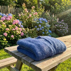 K&ouml;nnte beinhalten: Ein gefalteter, blauer Pullover liegt auf einem Picknicktisch aus Holz. Der Pullover hat eine weiche, strukturierte Oberfl&auml;che. Im Hintergrund ein Garten mit rosa und blauen Hortensien, einem Holzzaun und Gr&uuml;n, im Sonnenlicht.