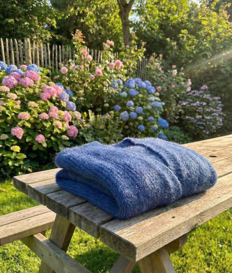 K&ouml;nnte beinhalten: Ein gefalteter, blauer Pullover liegt auf einem Picknicktisch aus Holz. Der Pullover hat eine weiche, strukturierte Oberfl&auml;che. Im Hintergrund ein Garten mit rosa und blauen Hortensien, einem Holzzaun und Gr&uuml;n, im Sonnenlicht.