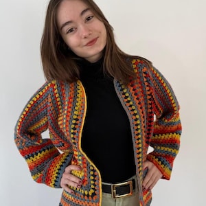 May include: A vibrant, hand-crocheted jacket with a geometric design. The jacket showcases a blend of orange, yellow, grey, and blue squares. It has long sleeves and a collarless style. The jacket is worn over a black polo neck and paired with a black belt and khaki trousers.