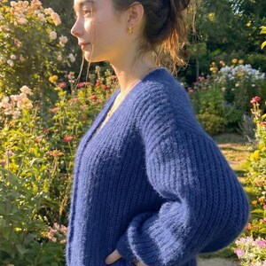 K&ouml;nnte beinhalten: Ein marineblauer, grob gestrickter Cardigan mit V-Ausschnitt. Der Pullover hat lange &Auml;rmel und wird in einem Garten mit Blumen im Hintergrund getragen. Der Cardigan wirkt weich und gem&uuml;tlich, ideal zum &Uuml;berziehen.