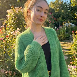 May include: A woman wearing a bright green, open-front cardigan. The cardigan is made of a fuzzy knit material. She is wearing a black top and black pants. The background is a garden with flowers and trees.