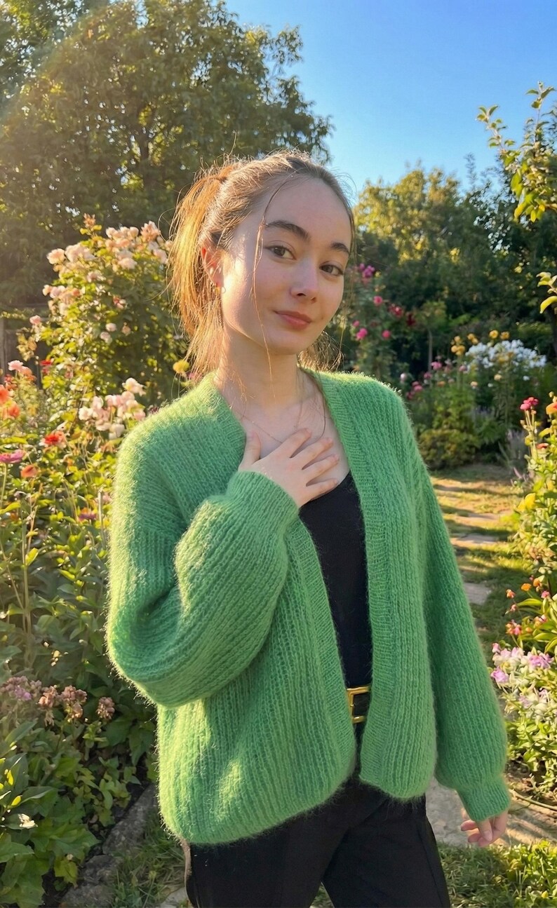 May include: A woman wearing a bright green, open-front cardigan. The cardigan is made of a fuzzy knit material. She is wearing a black top and black pants. The background is a garden with flowers and trees.