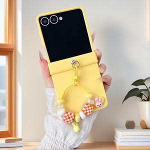 May include: A vibrant yellow phone case with a black screen, and a decorative charm. The charm includes a pink bunny, checkered beads, and yellow accents. The case is designed to fold open and closed.