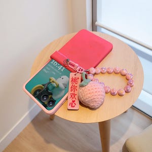 May include: A pink phone case with a phone displaying a cartoon image of a duck riding a bicycle. A pink beaded wrist strap and a pink knitted strawberry charm with a wooden tag that has Chinese characters are also present.