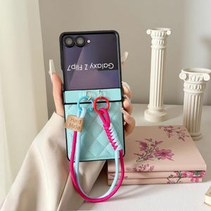 May include: A light blue quilted phone case with a black frame, designed for a Galaxy Z Flip7. It features a braided strap in pink and blue, and a small tag that says "Hand Made".