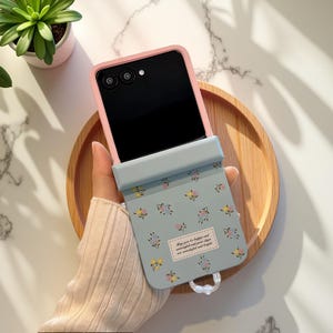 May include: A phone case with a light blue floral design. The case has a pink frame and a light blue flap with a floral pattern. The flap has a small label with text. The phone is black.