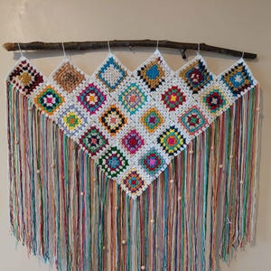 May include: A colorful, handmade wall hanging featuring a crocheted design with square motifs in various colors. The piece is suspended from a wooden branch and has long, multicolored fringe with wooden beads. A unique, bohemian-style home decor item.