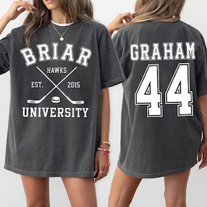 Könnte beinhalten: Dunkelgraues T-Shirt mit weißem Text und Grafiken. Auf der Vorderseite steht "Briar Hawks University" mit gekreuzten Hockeyschlägern und einem Puck. Auf der Rückseite stehen "Graham" und die Nummer "44" in Weiß.