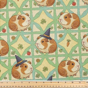 May include: A patterned fabric featuring cartoon guinea pigs in a repeating design. The guinea pigs are brown and white, with some wearing witch hats. The background has green, yellow, and blue geometric shapes, with apples, clovers, and carrots.