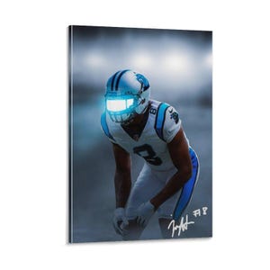 Puede incluir: Impresión en lienzo de un jugador de fútbol americano con el uniforme de los Carolina Panthers. El jugador está en posición agachada, con un casco con visera brillante. La imagen tiene una combinación de colores azul y blanco, con la firma del jugador y el número de camiseta visibles.
