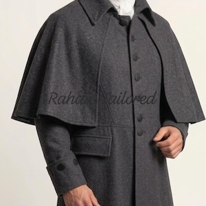 May include: A charcoal gray wool coat with a cape-like shoulder design. The coat features a notched collar, button closures, and a single pocket. The sleeves have buttoned cuffs. The text "Raha Tailored" is visible on the cape.