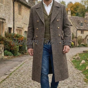May include: A brown herringbone double-breasted overcoat with gold buttons, worn over an olive green vest, a white collared shirt, and blue denim jeans. The coat features wide lapels and two side pockets. The text Raham Tailors is visible.