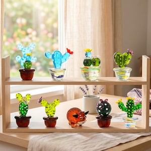 Könnte beinhalten: Ein Holzregal zeigt eine Sammlung farbenfroher Glas-Kaktus-Skulpturen. Die Kakteen variieren in Form und Farbe, darunter Blau, Grün und Gelb, und stehen in kleinen Töpfen. Ein dekorativer Glasbulle ist ebenfalls vorhanden.