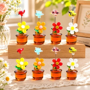 Könnte beinhalten: Ein Set aus acht Miniatur-Glasornamenten, die in zwei Reihen auf einem Holzblock angeordnet sind. Jedes Stück besteht aus einem kleinen bernsteinfarbenen Glastopf, einer bunten Glasblume und einem kleinen Tier- oder Insektenaufsatz aus Glas.