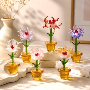 Könnte beinhalten: Sechs Miniatur-Glasblumenfiguren in bernsteinfarbenen Töpfen. Jede Figur hat einen grünen Stiel und eine einzigartige, mehrfarbige Blüte in Weiß, Rosa, Rot, Orange oder Violett. Die Blüten haben zarte, geschwungene Blätter und einen roten Kern.