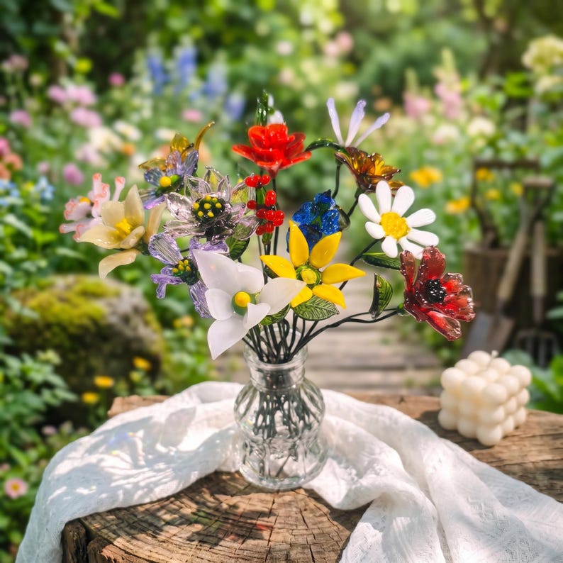 Puede incluir: Un colorido ramo de flores de cristal en un jarr&oacute;n de cristal transparente, colocado sobre una superficie de madera con un pa&ntilde;o blanco. El ramo presenta flores rojas, amarillas, blancas y azules, con tallos y hojas verdes. El fondo es un jard&iacute;n borroso.