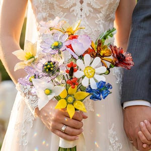 Puede incluir: Un colorido ramo de flores de cristal, que incluye margaritas, girasoles y otras flores en tonos amarillos, blancos, azules, rojos y morados. El ramo lo sostiene una persona con un vestido de novia de encaje blanco, con una persona con un traje gris.