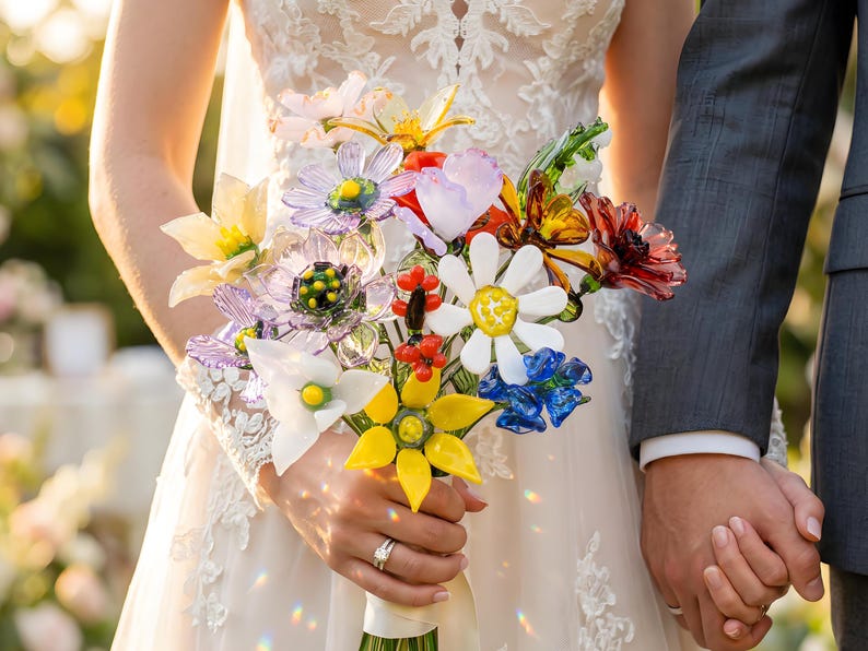 Puede incluir: Un colorido ramo de flores de cristal, que incluye margaritas, girasoles y otras flores en tonos amarillos, blancos, azules, rojos y morados. El ramo lo sostiene una persona con un vestido de novia de encaje blanco, con una persona con un traje gris.